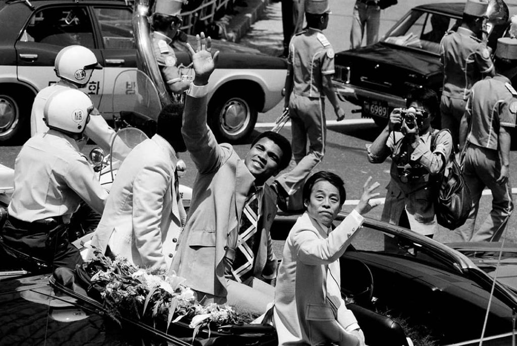 Muhammad Ali sitting on top of a car n-between two unknown men. Seoul, Korea (1976)